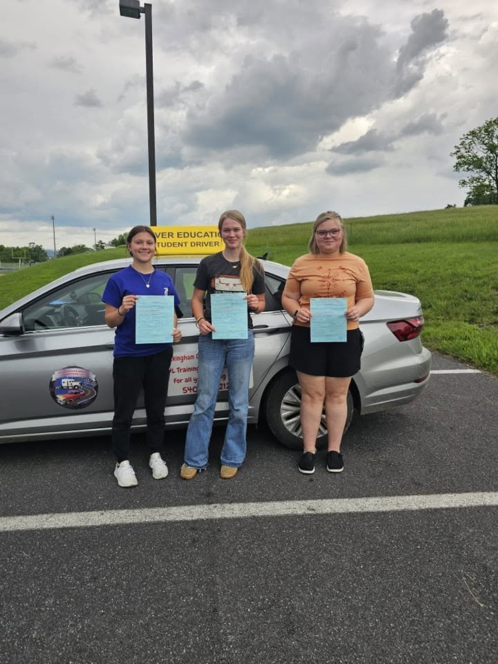 Three young women standing in front of a silver car with a learner driver sign on the roof, each holding a certificate or document. They are outdoors on a cloudy day, on a paved parking lot with a grassy area behind them.
