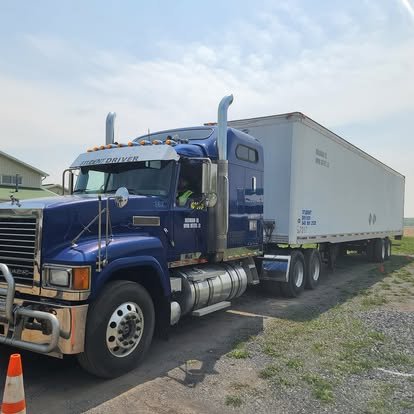 A blue semi-truck with a white trailer parked on a gravel lot near a grassy area and a house.