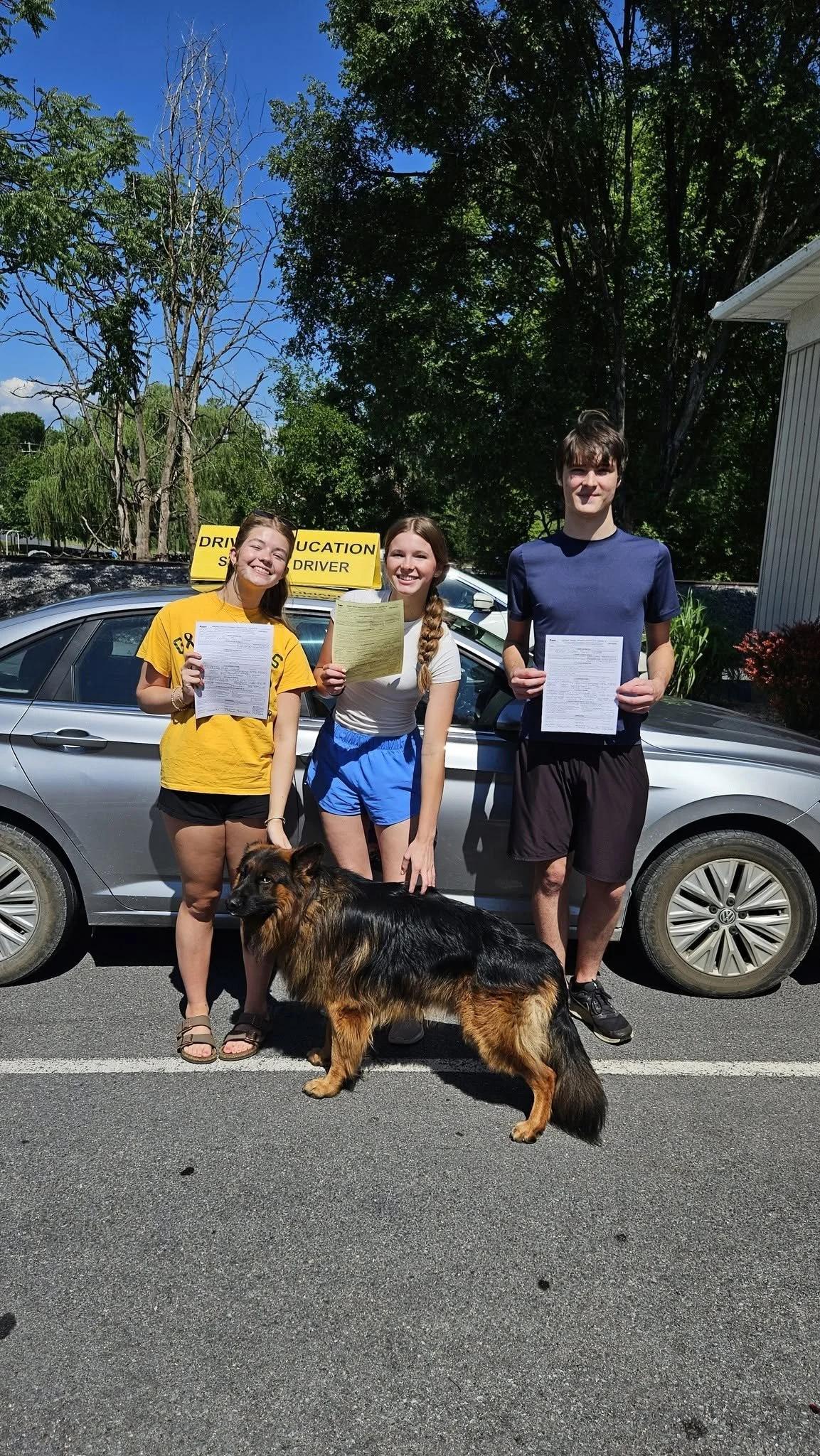 Group of three teenagers holding driver's licenses and documents, standing in front of a silver car with a yellow 'DRIVER EDUCATION' sign on top. A German Shepherd dog is in front of them, and trees and a building are in the background.