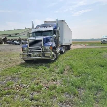 Blue semi-truck parked on grassy area near a large building with other vehicles in the background.