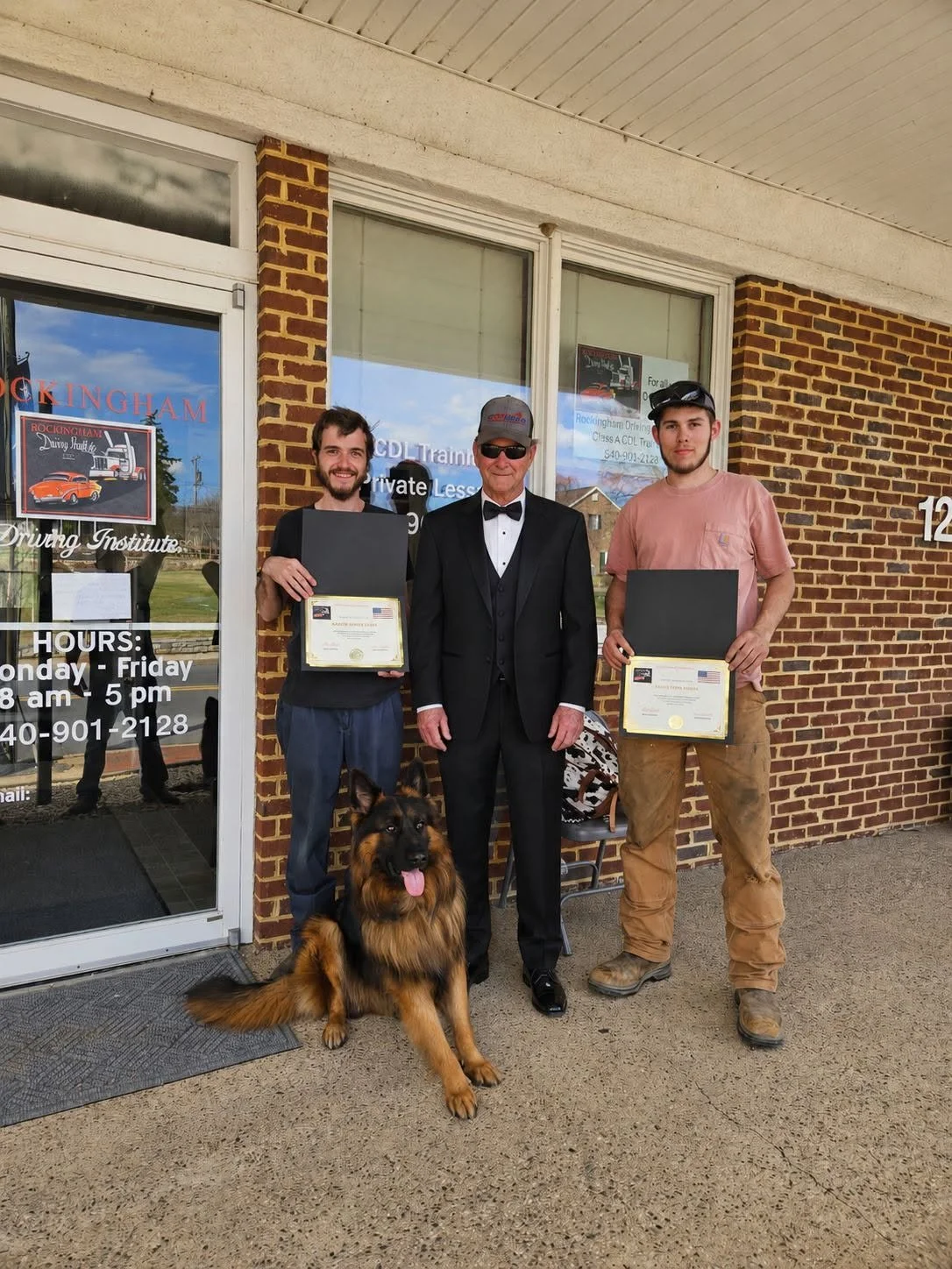 Two young men and an older man dressed in a tuxedo standing outside a brick building, each holding a Certificate of Completion, with a German Shepherd sitting in front of them. The building has glass doors and windows with signage, and the outdoor clock shows daylight.