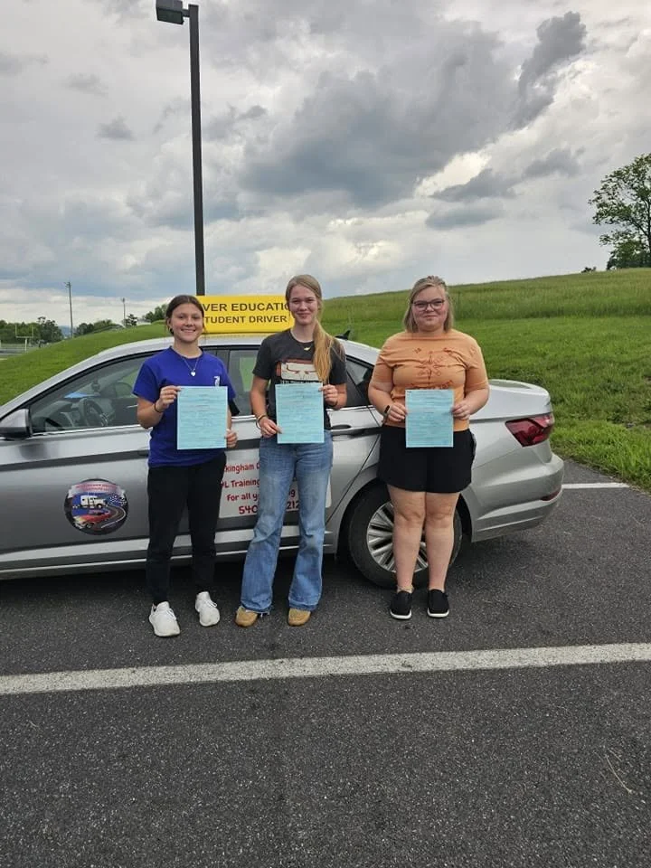 Three young women standing in front of a silver car holding certificates, under a cloudy sky in a parking lot.