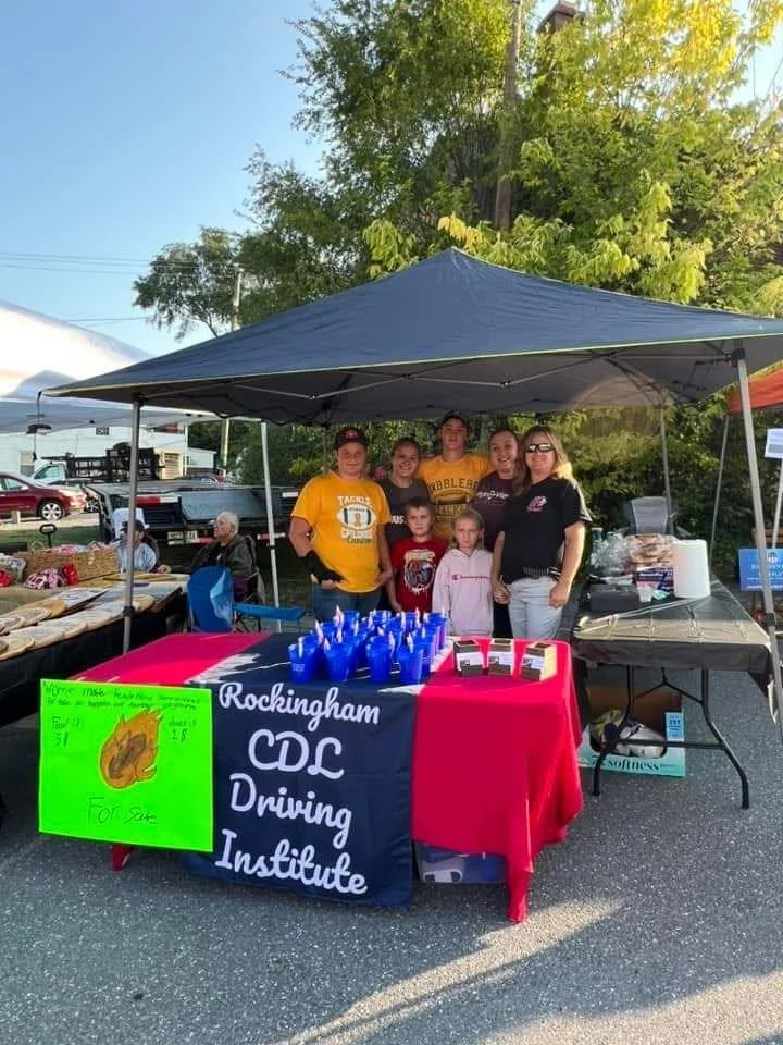 Group of seven people, including children, standing under a blue canopy at a community event, with a table displaying blue water bottles and flyers in front of them, and a sign for Rockingham CDC Driving Institute on the table.