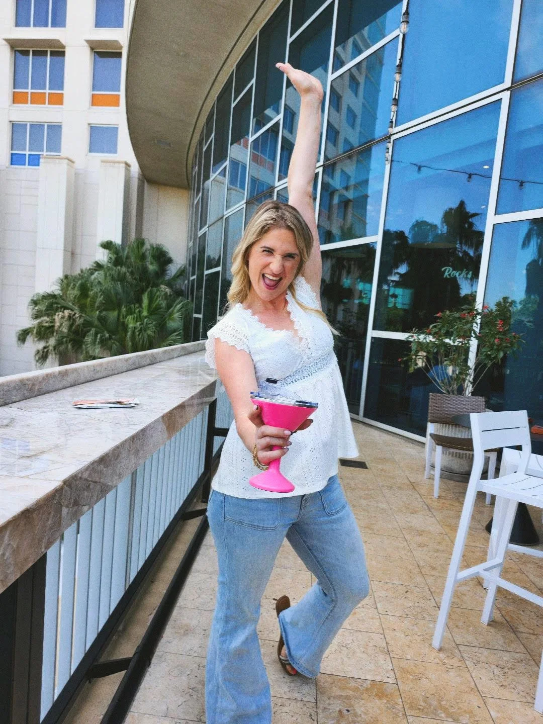 A woman standing outdoors, holding a pink cocktail glass, wearing a white lacy top and blue jeans, and posing with her arm raised and a playful expression.