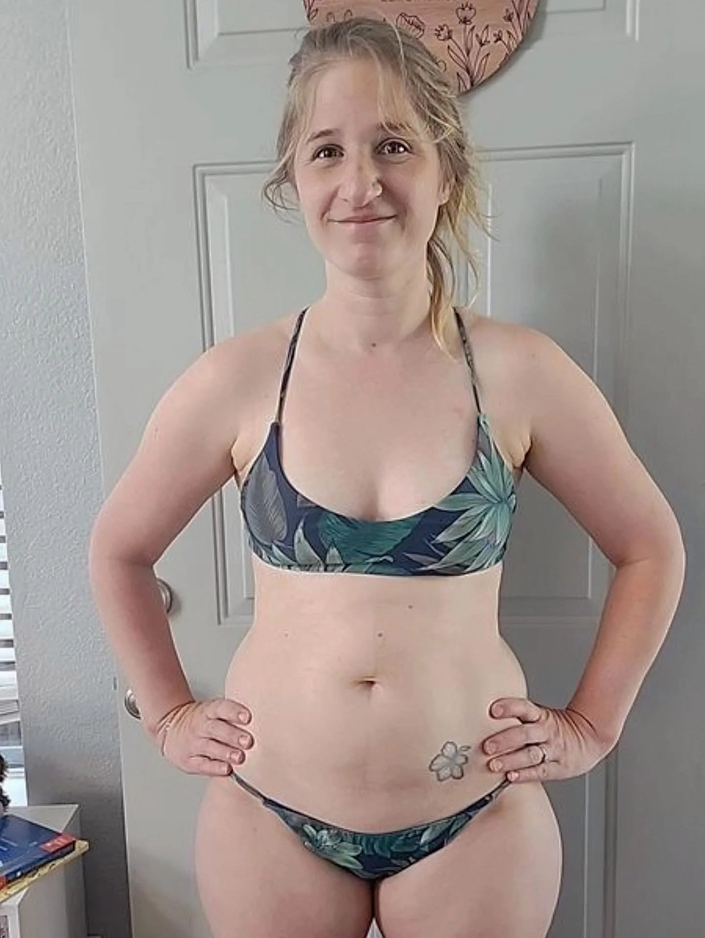 A woman with blonde hair, standing with her hands on her hips, wearing a tropical-patterned bikini top and bottoms, standing in front of a gray door.