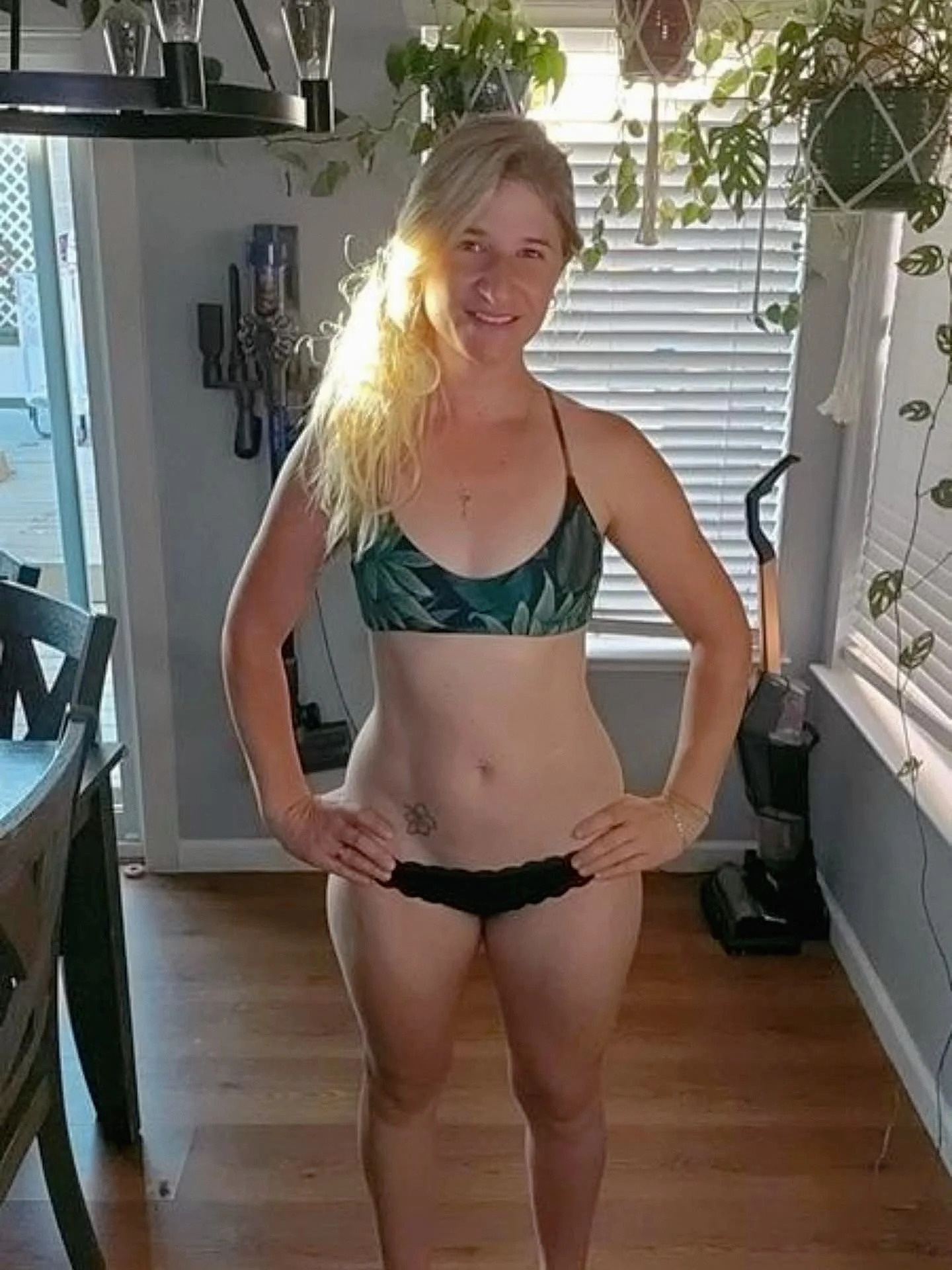 A woman in a leafy-patterned bikini top and black underwear, standing in a bright room with plants and blinds, smiling with her hands on her hips.