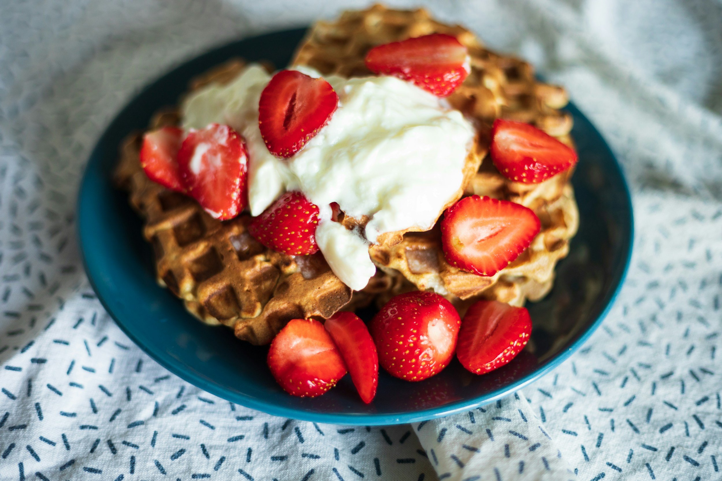 Wafels met slagroom en aardbeien op een blauw bord, op een wit tafellaken.
