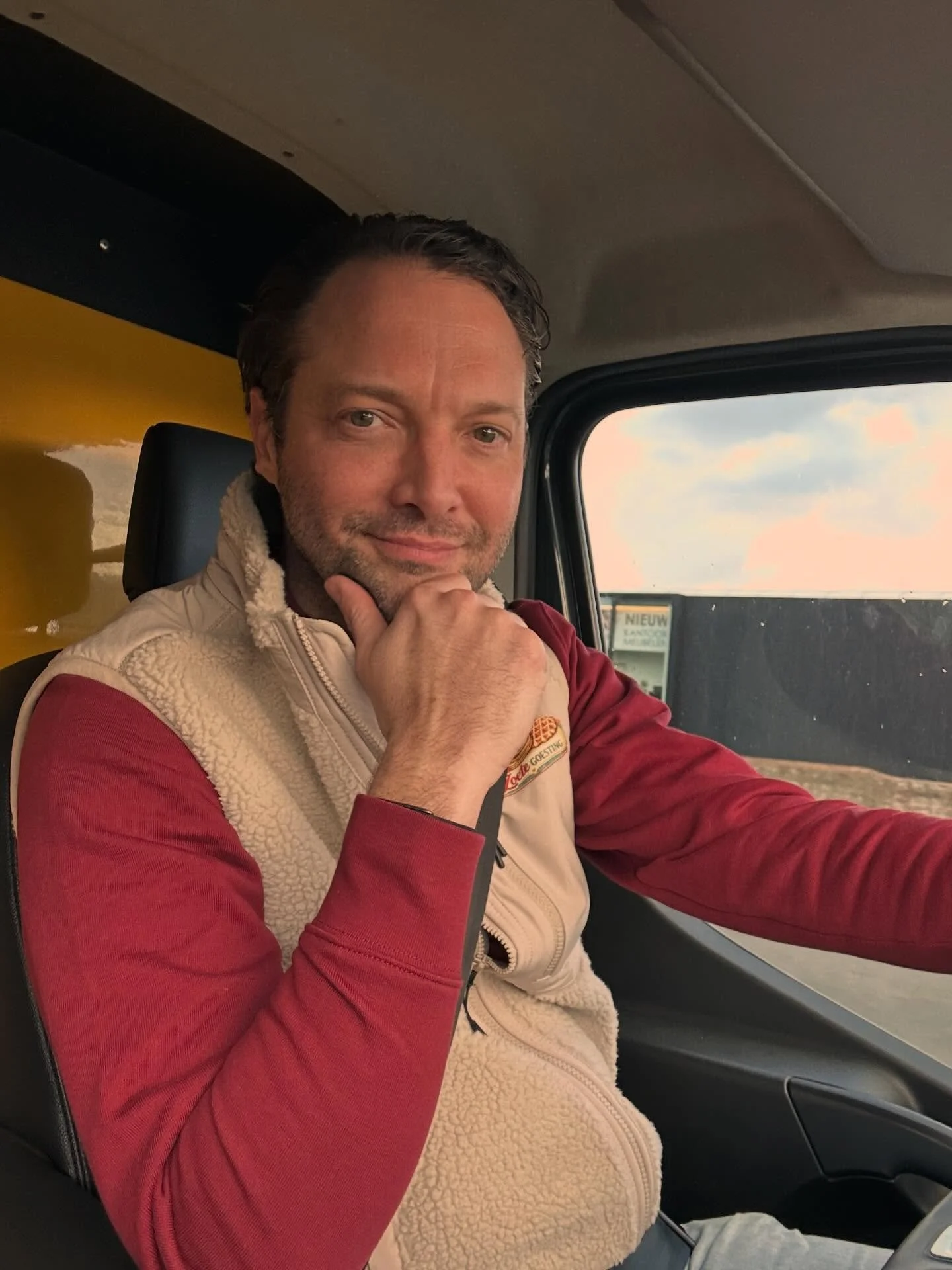 Man zit in de bestuurdersstoel van een voertuig, met de hand onder de kin, terwijl hij naar de camera kijkt, buiten het raam is een landschap zichtbaar.