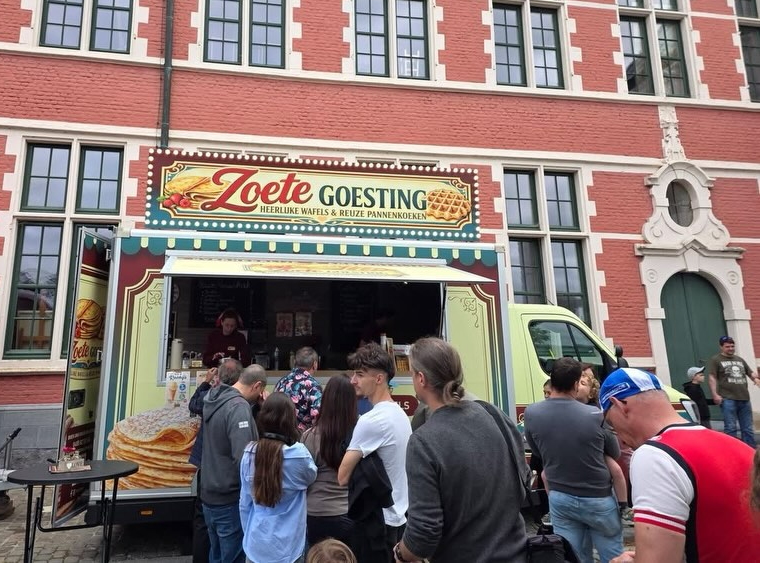 Een foodtruck genaamd 'Zoute Going' die verse wafels en pannenkoeken verkoopt, en een menigte mensen die in de rij staan om te bestellen, voor een rood-bruin gebouw.