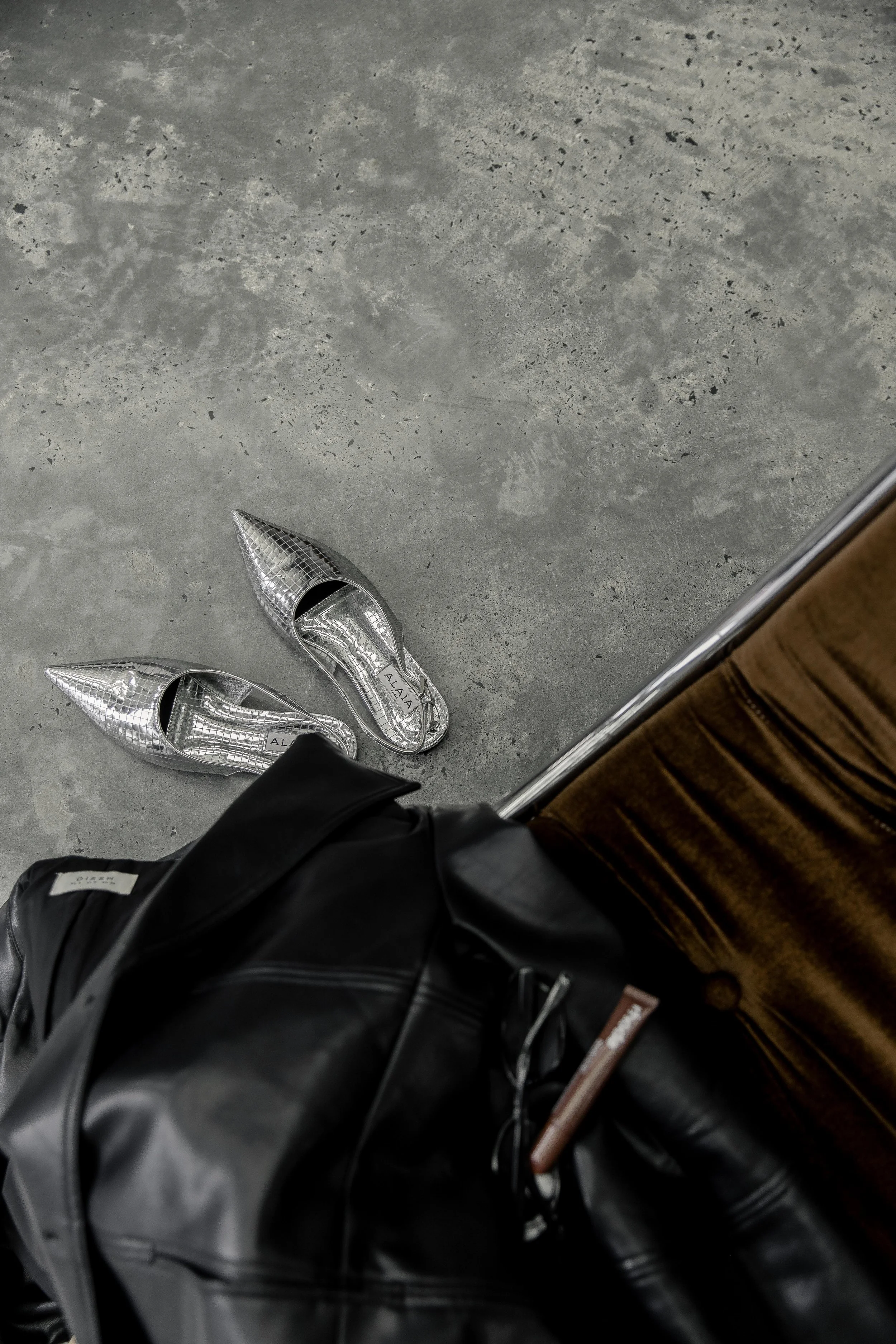 A pair of silver high-heeled shoes on a concrete floor, viewed from above. Part of a person’s black leather jacket and a leather bag are also visible.