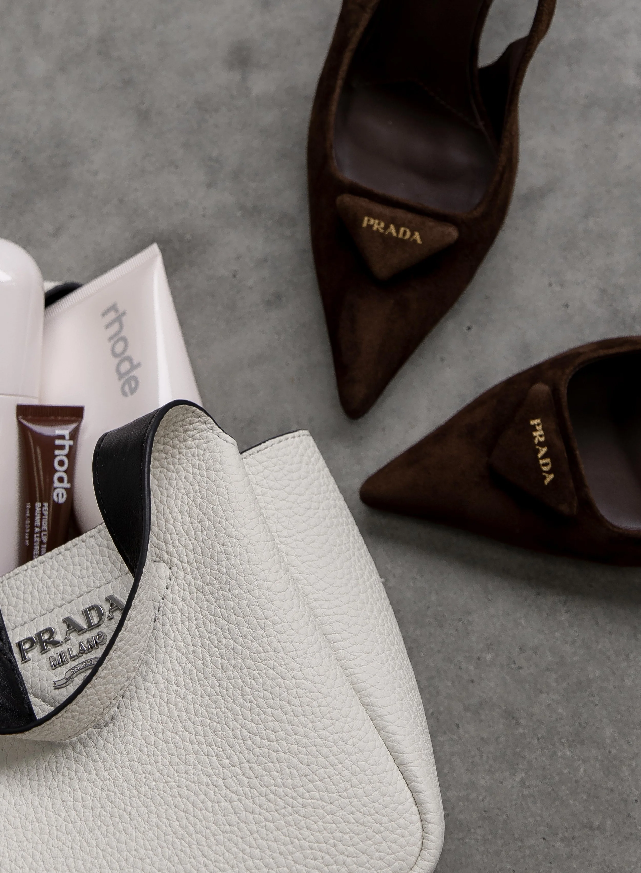 Close-up of a white Prada leather handbag, with a black leather handle and a Prada logo patch on the front, on a light gray surface. Two pairs of brown Prada high heels are nearby.