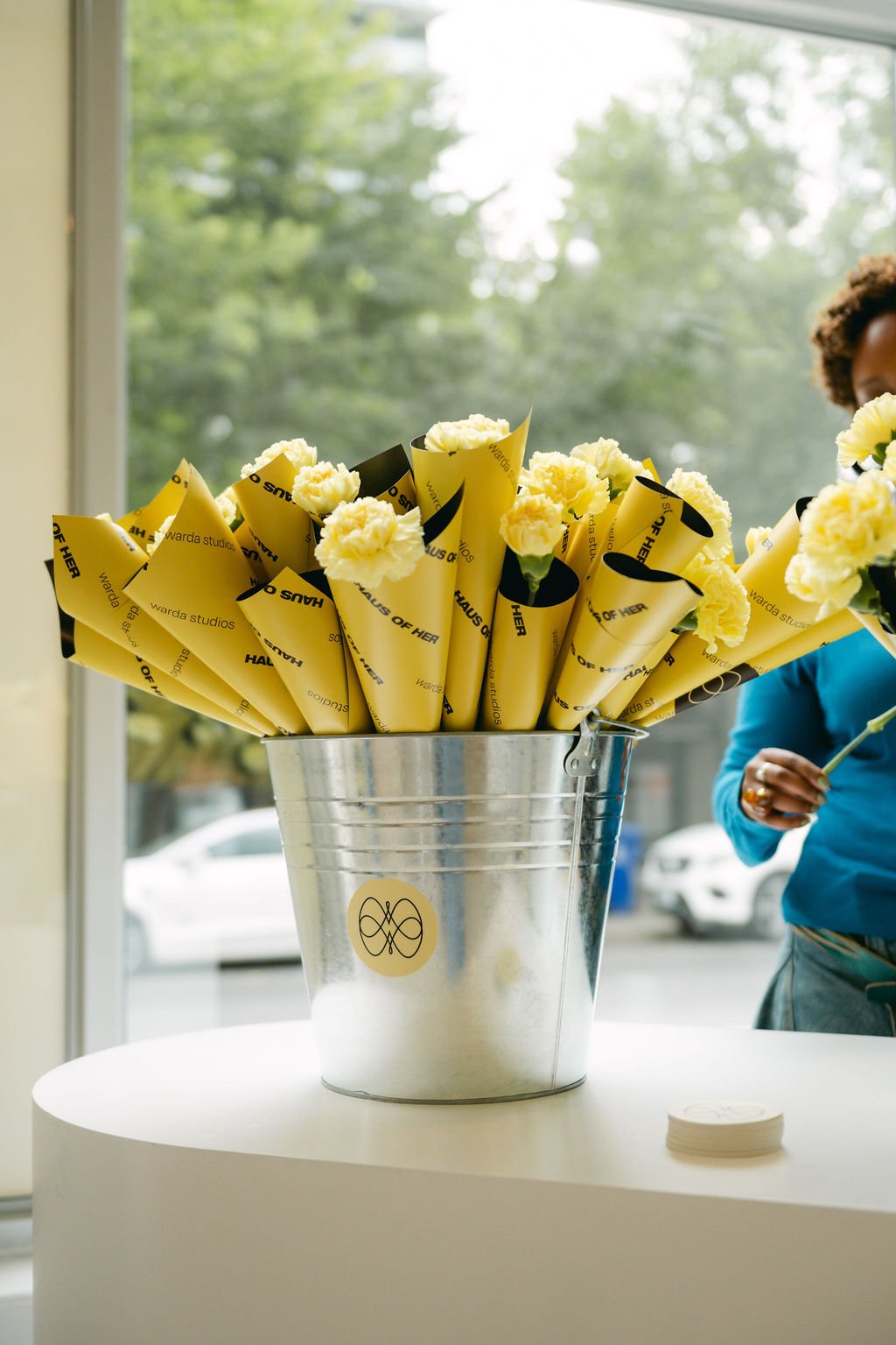 A metallic bucket filled with yellow flowers wrapped in yellow and black paper with the text 'Haus of Her' and 'Warda Studios' on it, placed on a white round table near a large window with trees outside.