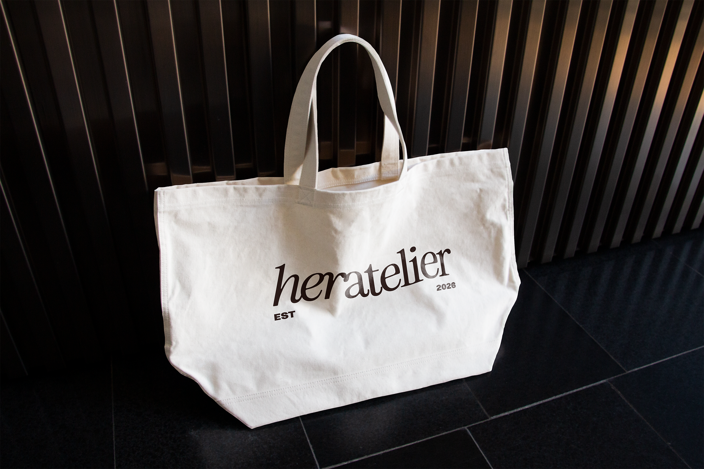A white tote bag hanging on a black slatted wall, with the text 'homteller' printed on it, and smaller text 'EST 2026' beneath.