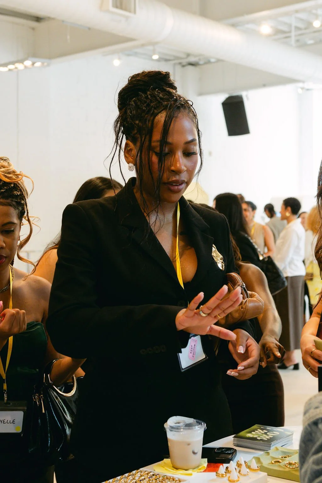 A woman with dark, braided hair styled in an updo wearing a black blazer, with a yellow lanyard, standing at a booth with various jewelry and an iced coffee, engaging with someone at an indoor event.