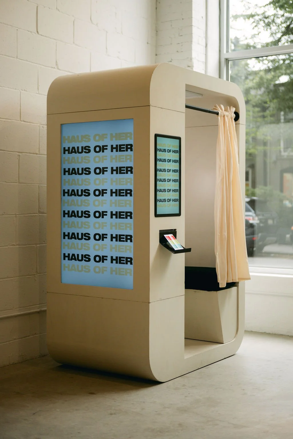 A modern photo booth with a large screen displaying the text 'HAUS OF HER' repeated in blue, black, and light green colors, situated in front of a white brick wall near a window with curtains.