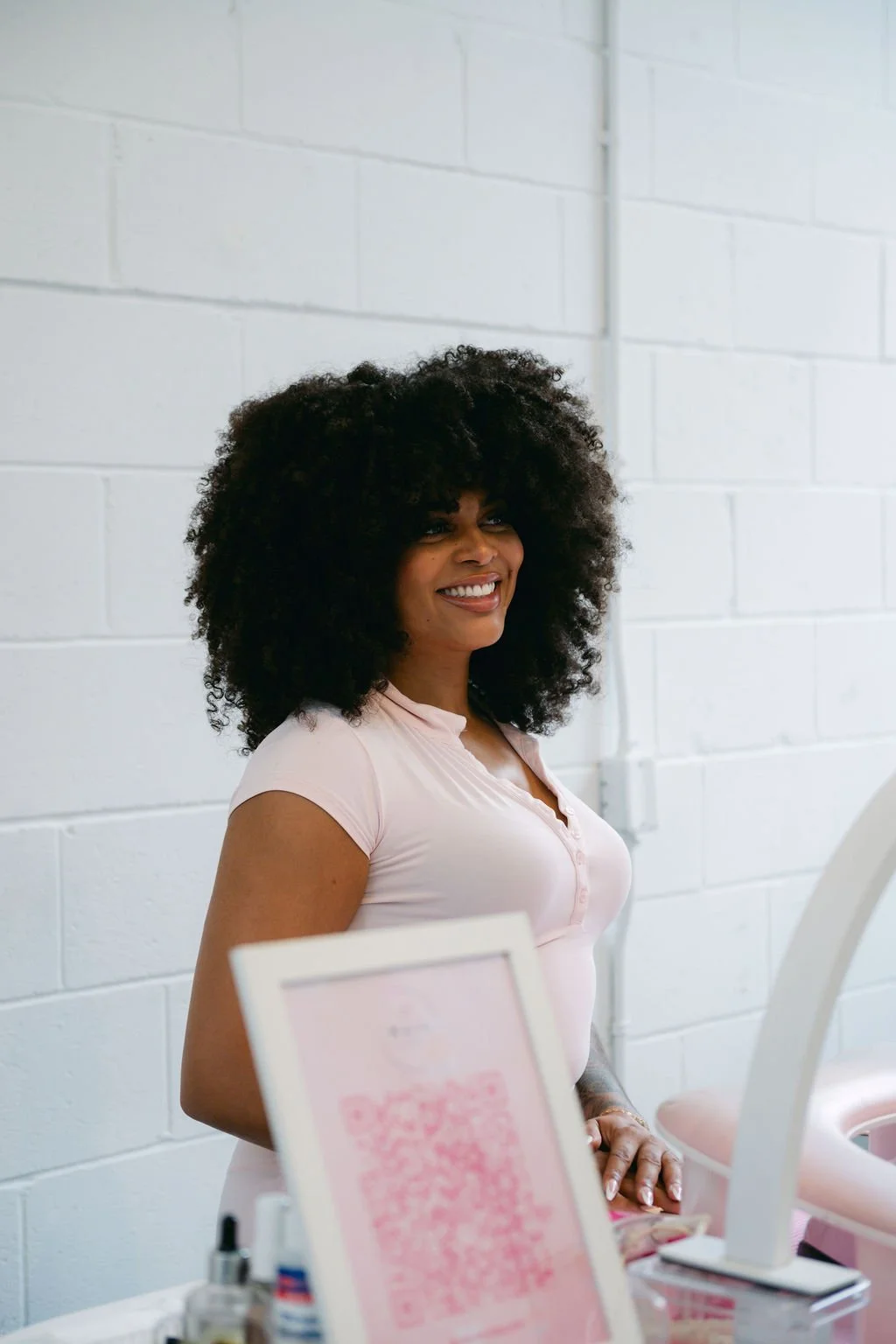 A woman with dark curly hair smiling, standing in a light-colored room with white brick walls, near a table with beauty products and a pink QR code poster.