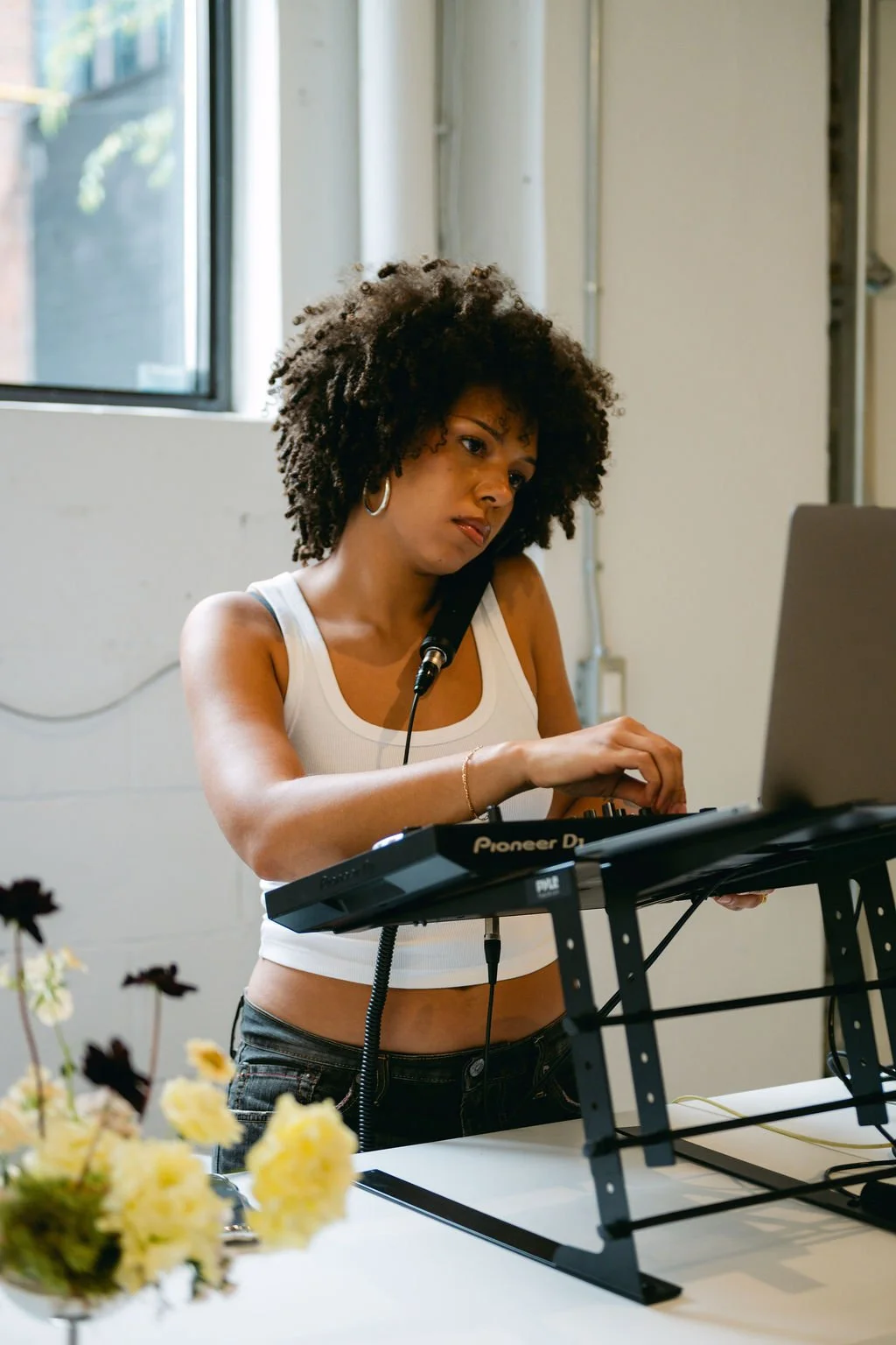 A woman with curly hair wearing a white tank top is working with music equipment, possibly DJing or producing music, in a well-lit room with a window.