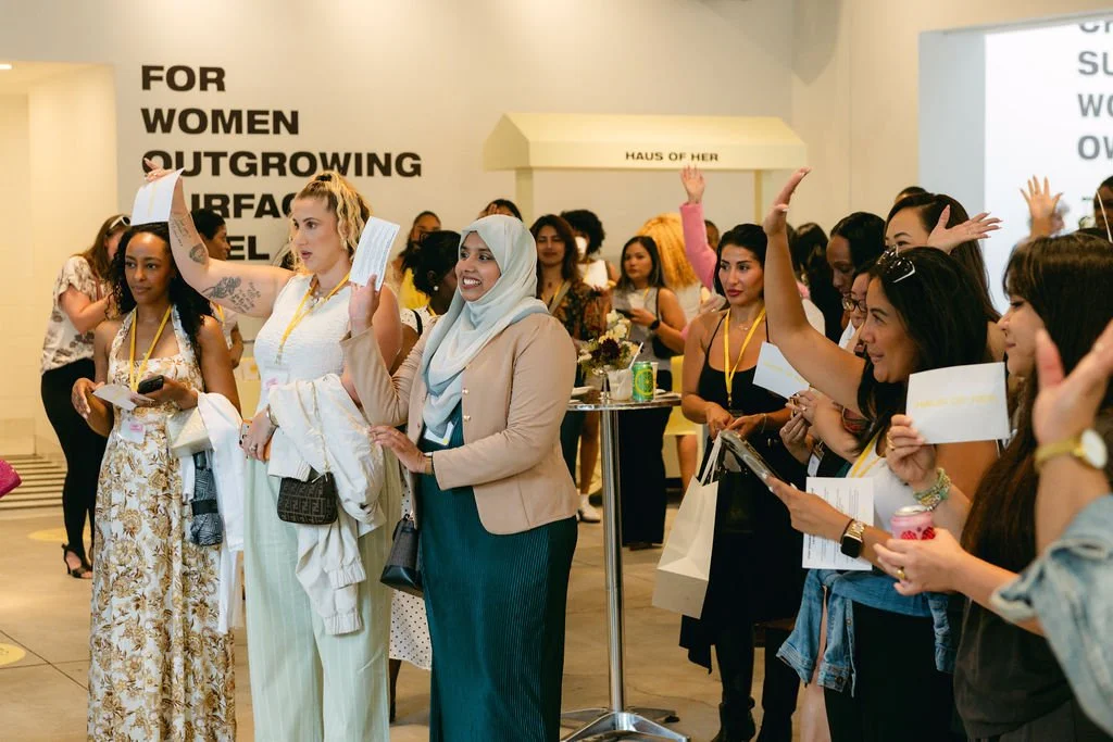 Group of women gathered indoors at an event, some raising their hands, holding papers and shopping bags, with a large sign on the wall behind them that reads 'FOR WOMEN OUTGROWING SURFACE LEVEL'.