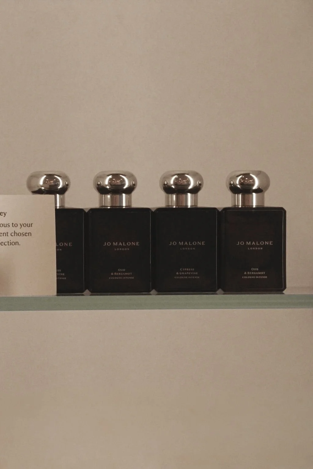 Four bottles of Jo Malone London cologne with silver ball-shaped caps, labeled Oud & Bergamot, Oud & Bergamot, Cypress & Grapevine, and Oud & Bergamot, placed on a shelf.