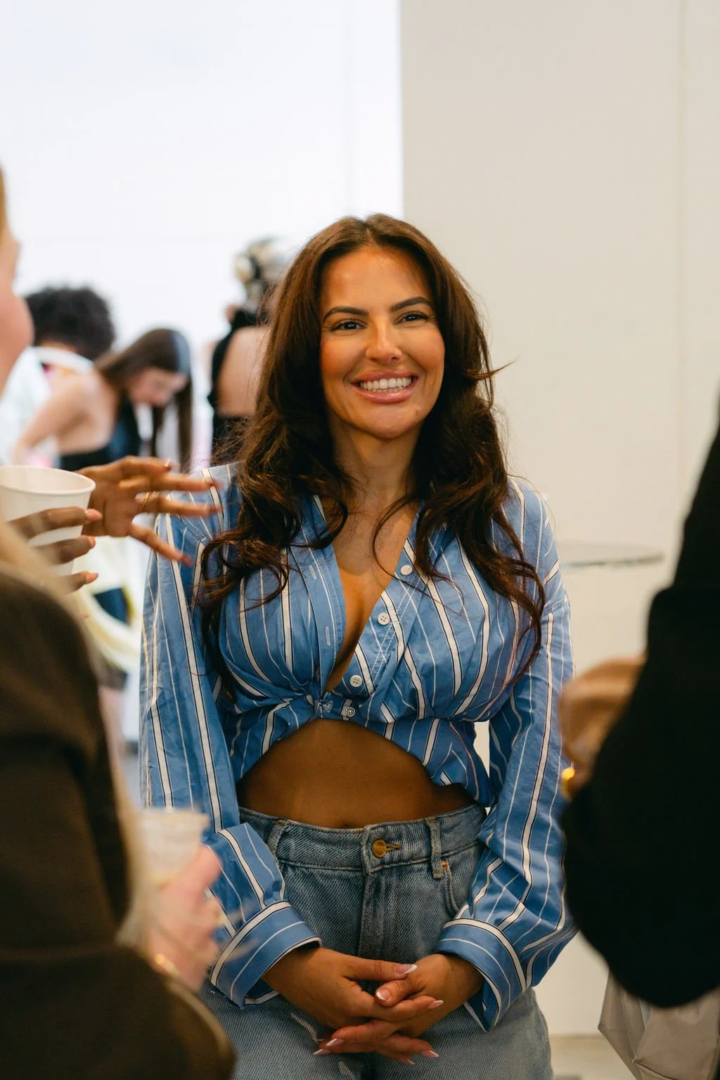 A woman with long dark hair, wearing a blue and white striped shirt with a midriff cutout and high-waisted jeans, smiling at a social gathering indoors.