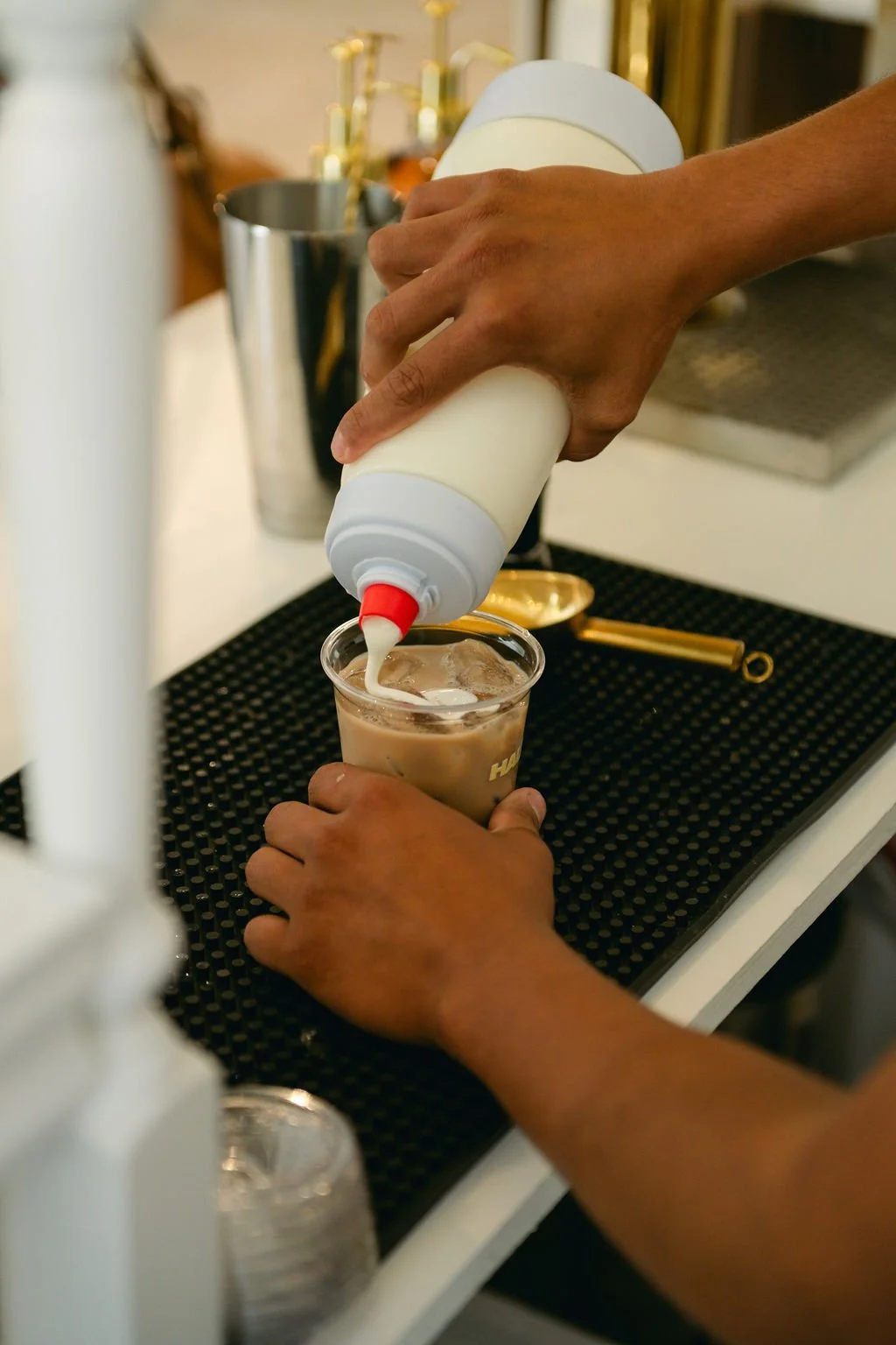 A person pouring milk into iced coffee in a clear cup at a bar or cafe counter.
