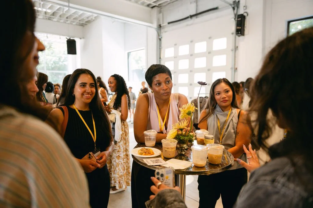 A group of women at a social gathering or event, standing around a high table with drinks and snacks in a bright, spacious room with white walls and garage-style doors.