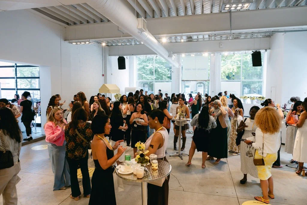 A crowded social event with women gathered and talking in a spacious, well-lit room with large windows.