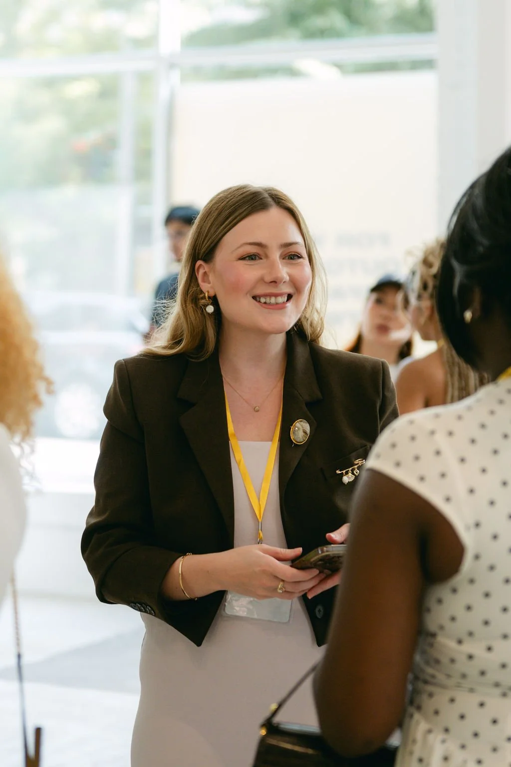 A woman with light brown hair and earrings smiling and talking to another woman with dark skin, wearing a white polka dot dress, at a professional event inside a bright room.