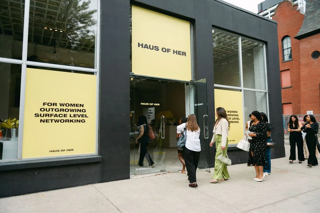 People entering and standing outside the storefront of Haus of Her, a retail store with large yellow signs promoting women and surface level networking.
