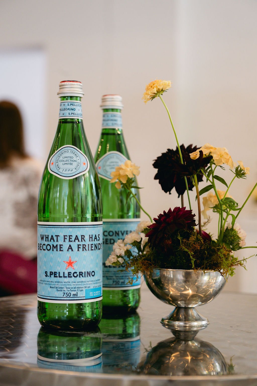 Two green bottles of S. Pellegrino sparkling water with custom labels on a reflective surface, accompanied by a silver bowl filled with white and dark red flowers, set against a plain background.