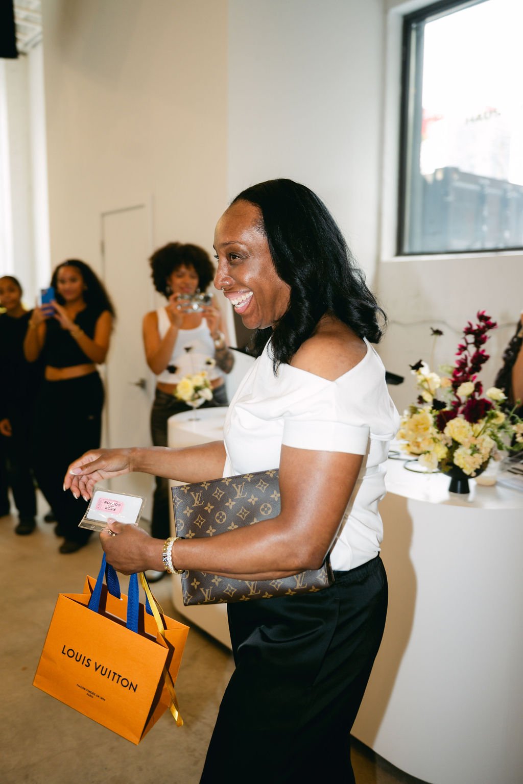 A woman smiling at an indoor event, holding a Louis Vuitton shopping bag and a designer clutch. In the background, several women are taking photos and holding flowers, with a table of flowers nearby.