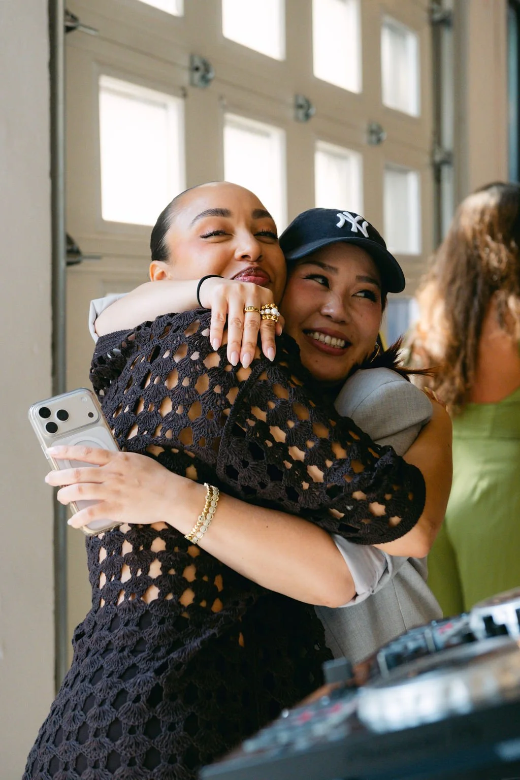 Two women hugging, smiling, and celebrating together indoors during the daytime. One woman is holding a smartphone, and they appear to be joyful.