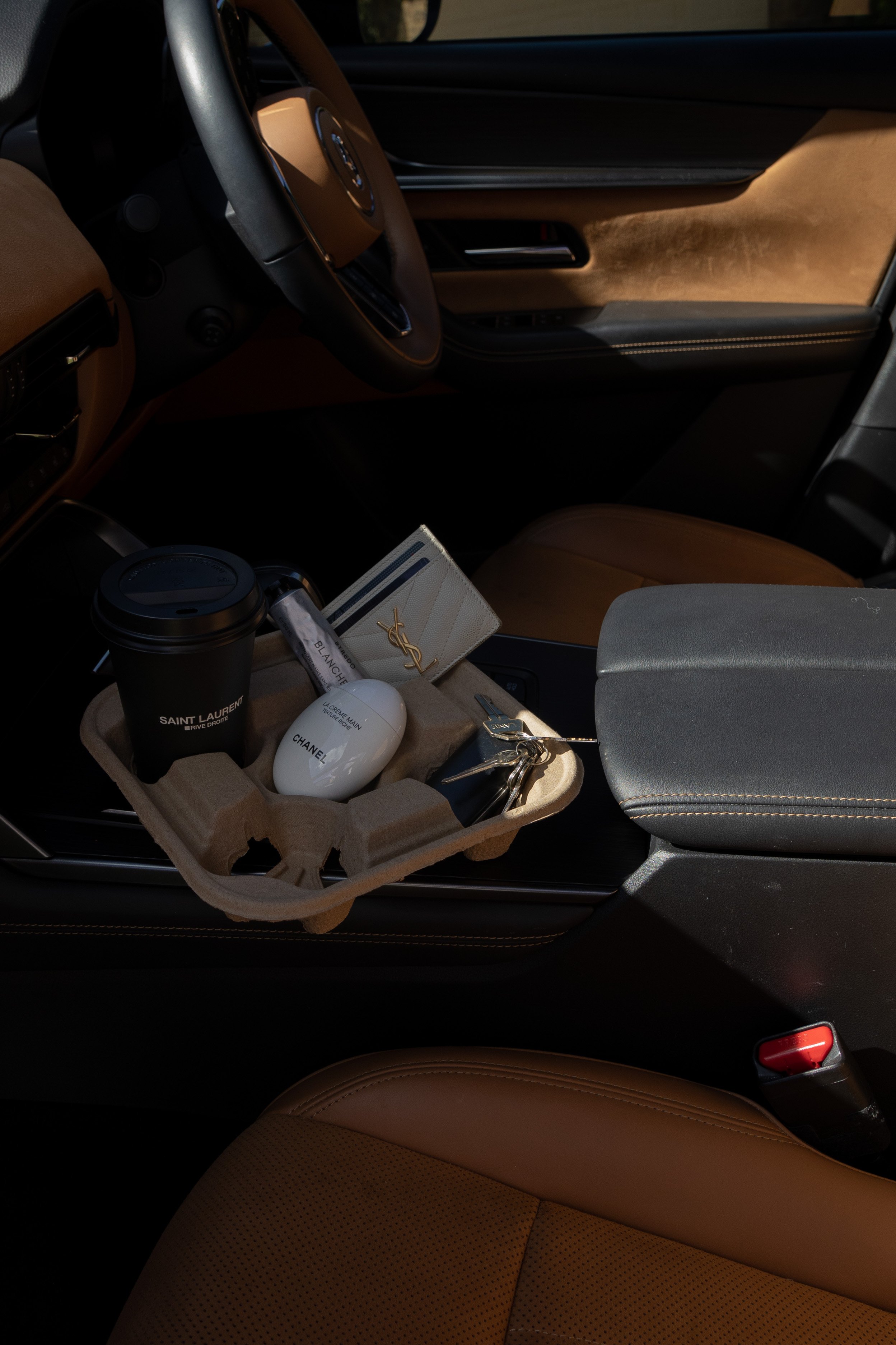 The interior of a luxury car with tan leather seats, a black steering wheel, and a center console holding a Saint Laurent branded beverage cup, a Chanel perfume bottle, and a set of keys on a beige tray.