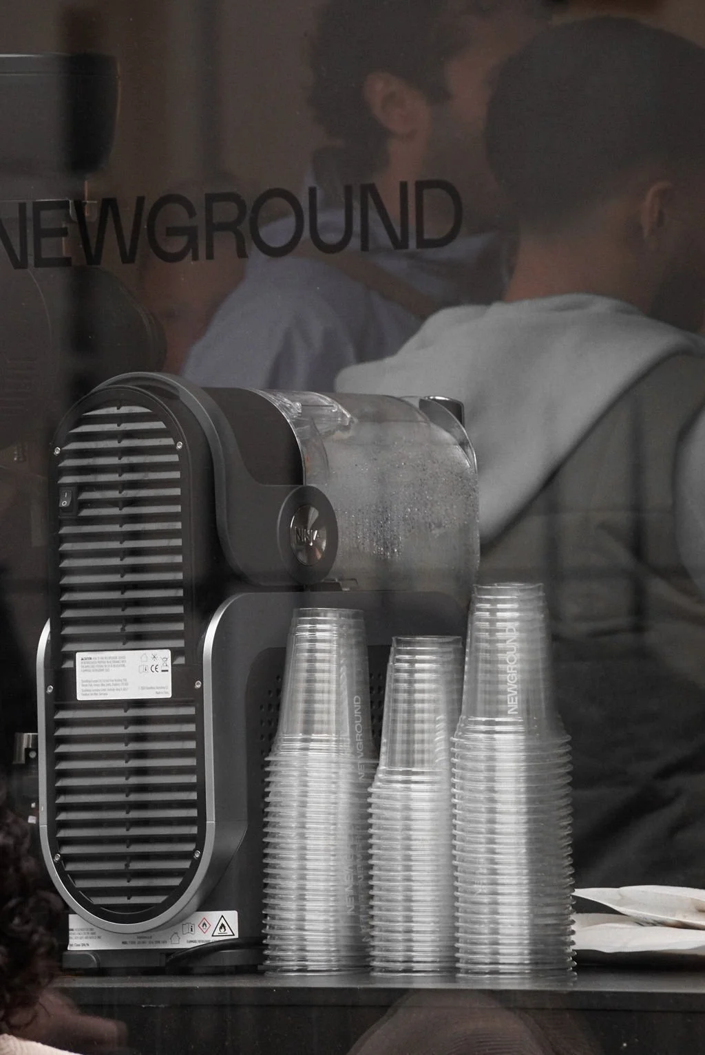 Stack of clear plastic cups next to a coffee machine with someone visible through a glass wall that has the words 'NEW GROUND' printed on it.