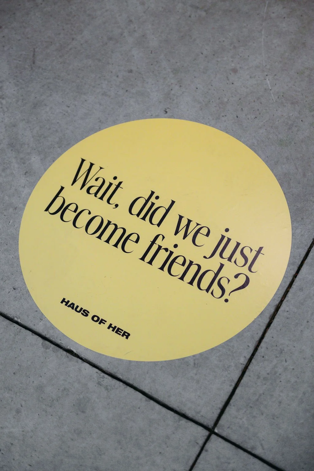 A yellow circular sticker on the floor with the text "Wait, did we just become friends?" and "HAUS OF HER" written below.