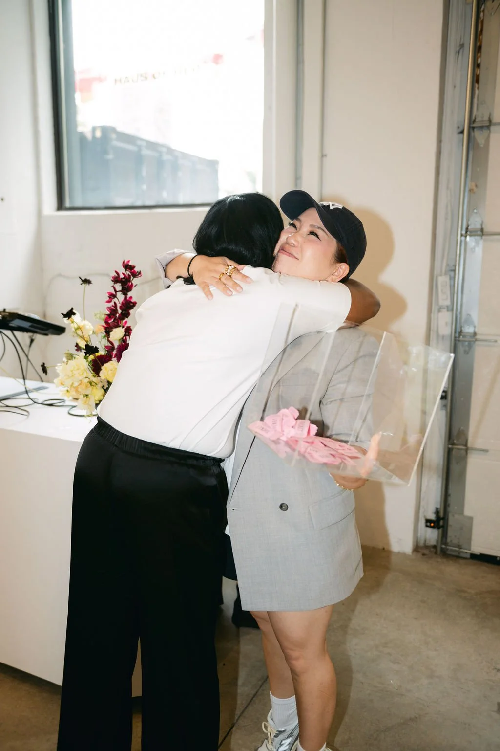 Two women embrace in a hug, one woman wearing a black cap and holding a pink gift, the other woman with shoulder-length black hair. The scene is indoors with a window in the background and flowers on a table.