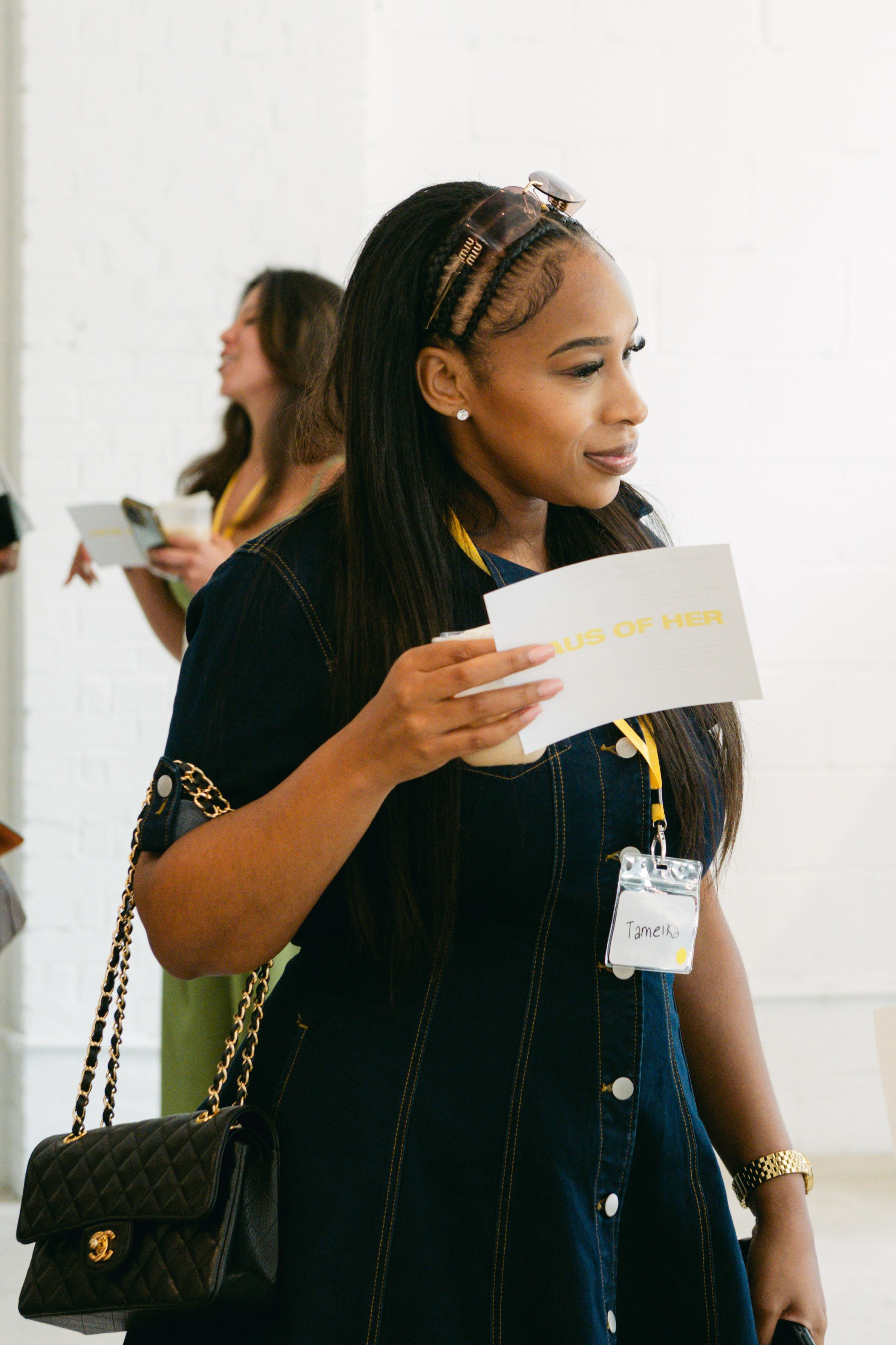 A woman with long black hair, wearing glasses on her head and a dark denim dress, holds a paper and a cellphone, standing in a brightly lit room with a white brick wall. Another woman with brunette hair and a yellow strap top is in the background, sm