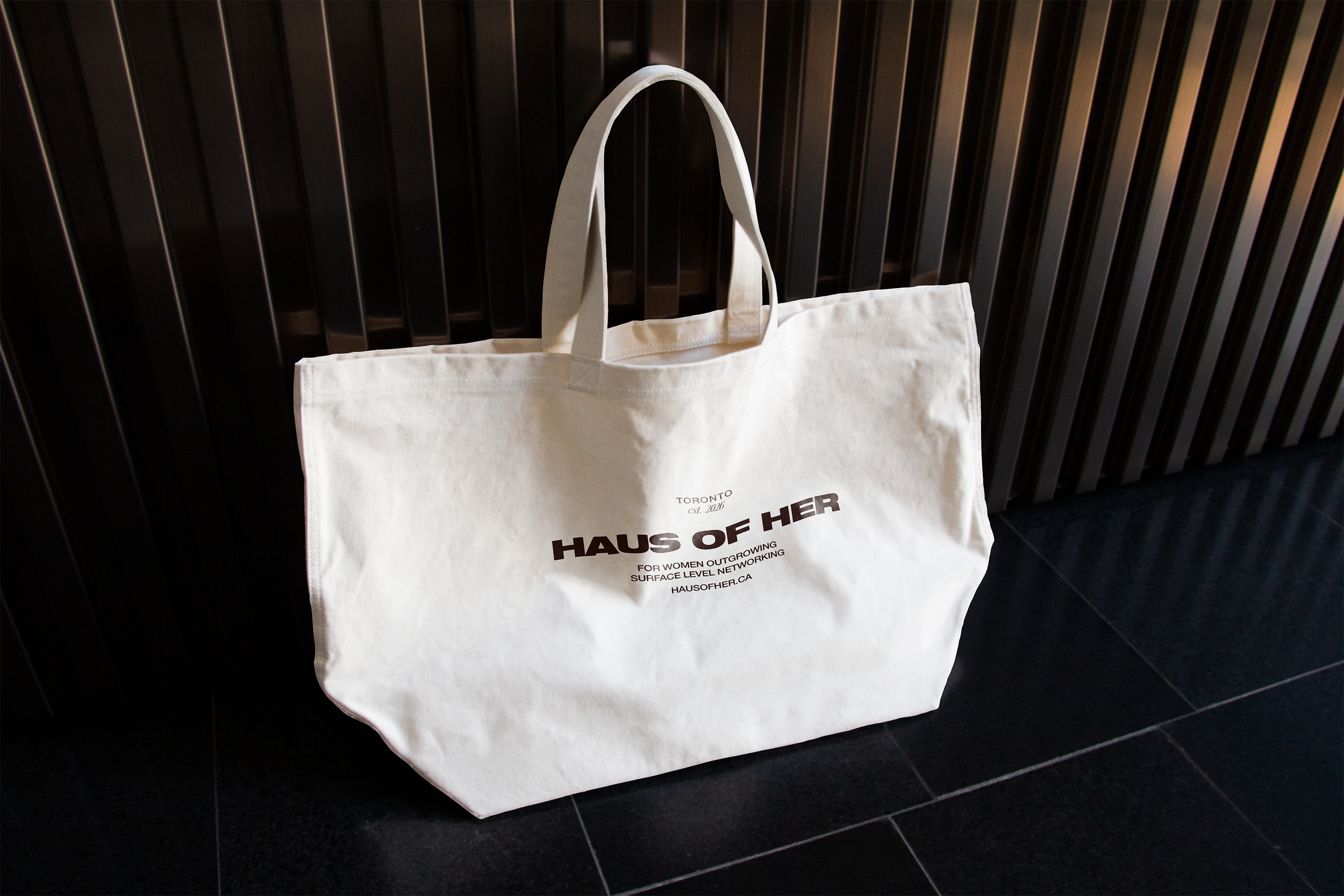 A white tote bag hanging on a black slat wall. The tote bag has black text promoting HAUS OF HER, a network for women in surface-level technology, based in Toronto, est. 2016.