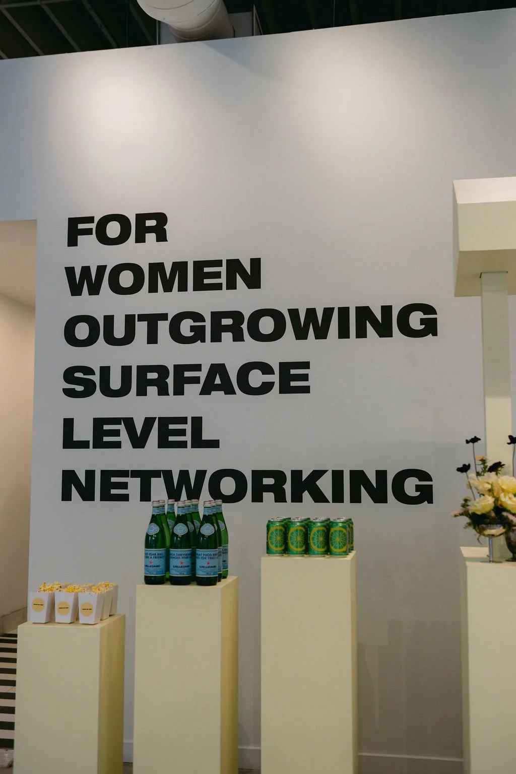 Large wall sign reading 'For Women Outgrowing Surface Level Networking' with display tables holding bottled beverages, canned drinks, and a flower arrangement.