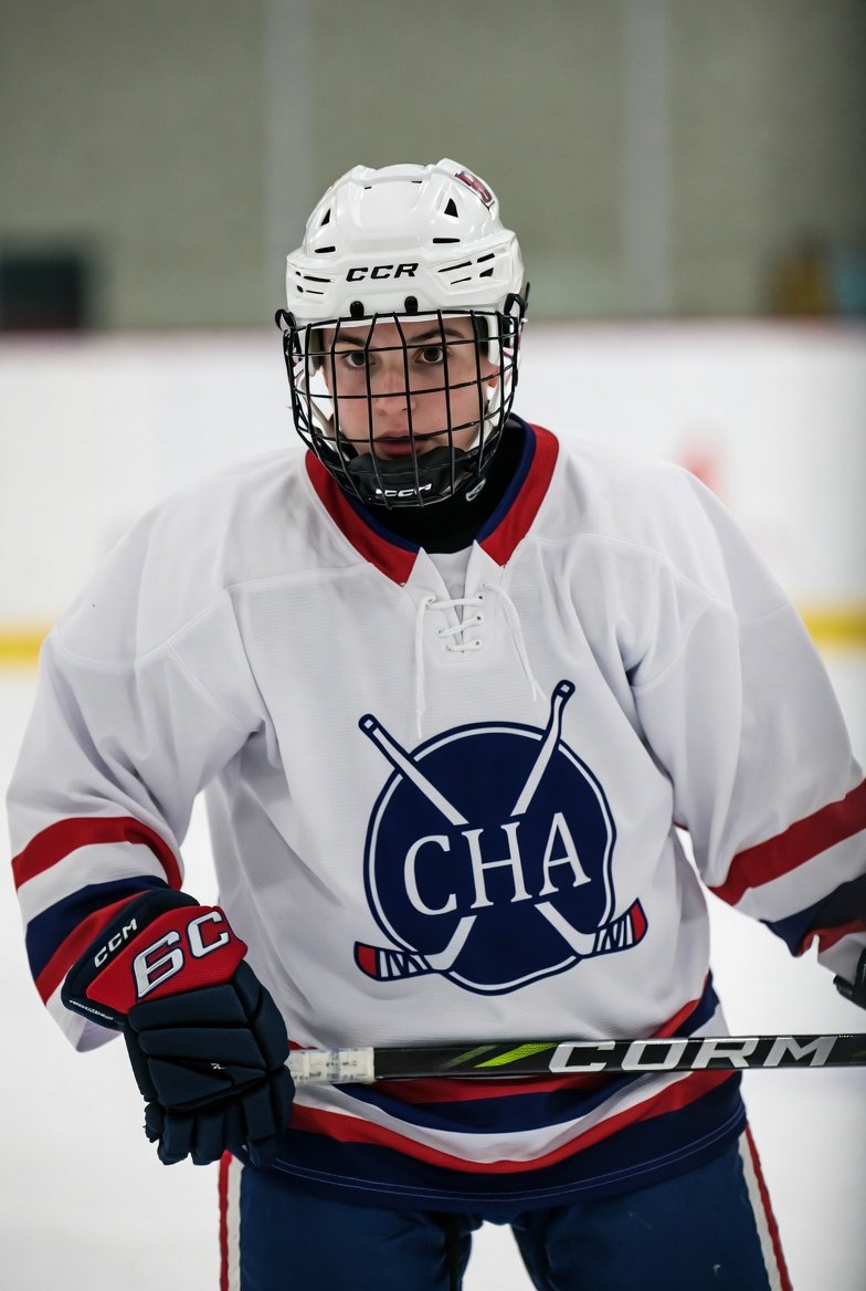 Young ice hockey player in full gear on the rink, wearing a white jersey with a logo of crossed hockey sticks and a puck, holding a hockey stick, and looking at the camera.