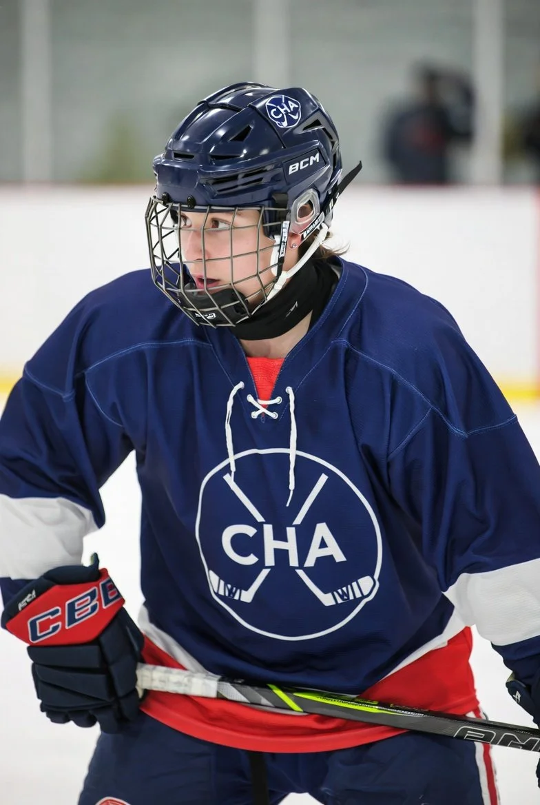 A person wearing a navy blue hockey helmet with cage, navy blue hockey jersey with white emblem and red pants, holding a black hockey stick on an ice rink.