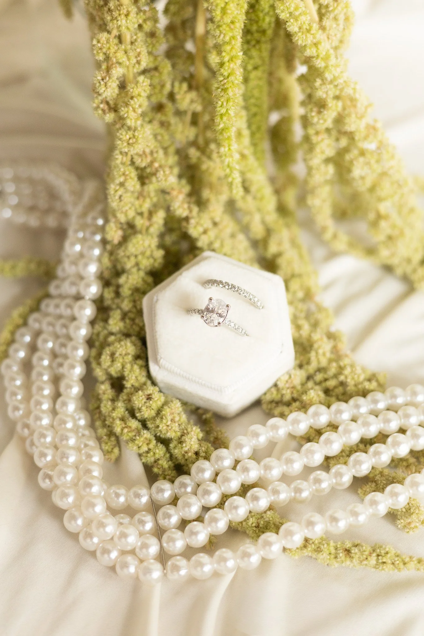 Close-up of elegant jewelry, including a diamond ring on a white ring box, surrounded by multiple strands of white pearls and cream-colored flowers.