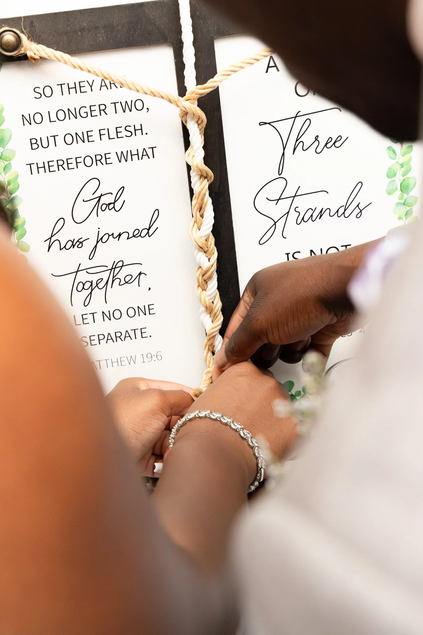 Close-up of hands holding a rope with a white and beige twisted pattern, against a backdrop of printed paper with text about God and unity. The person is wearing jewelry, including a diamond ring and a bracelet.