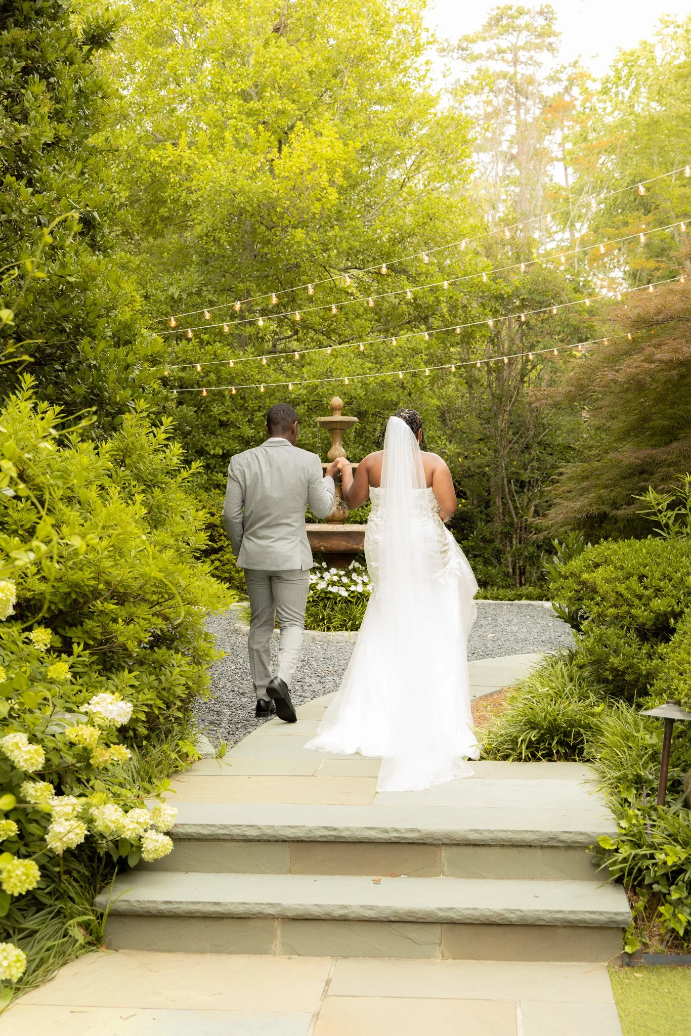 A bride and groom walking up steps in a garden, holding hands, with string lights hanging above them during daytime.