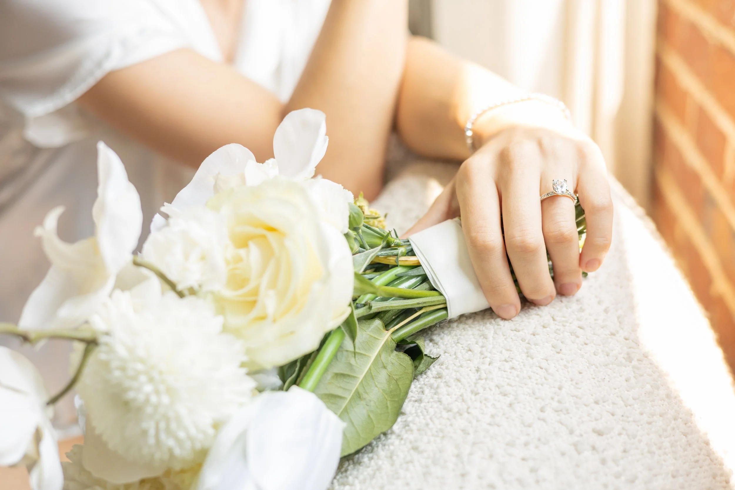 A bride's hand with a diamond engagement ring holding a bouquet of white flowers on a table covered with white material.