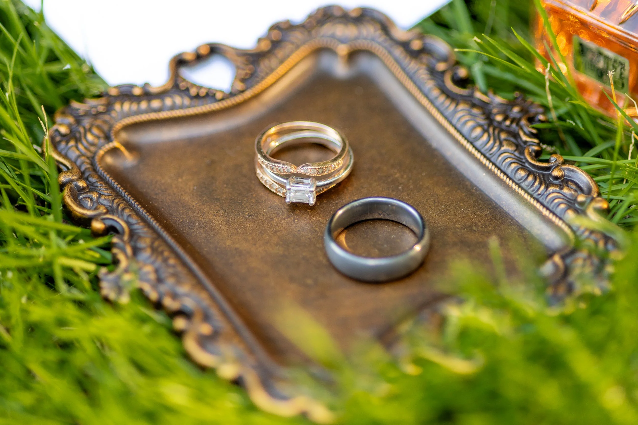 Close-up of engagement and wedding rings on an ornate silver tray, placed on green grass with a blurred background.