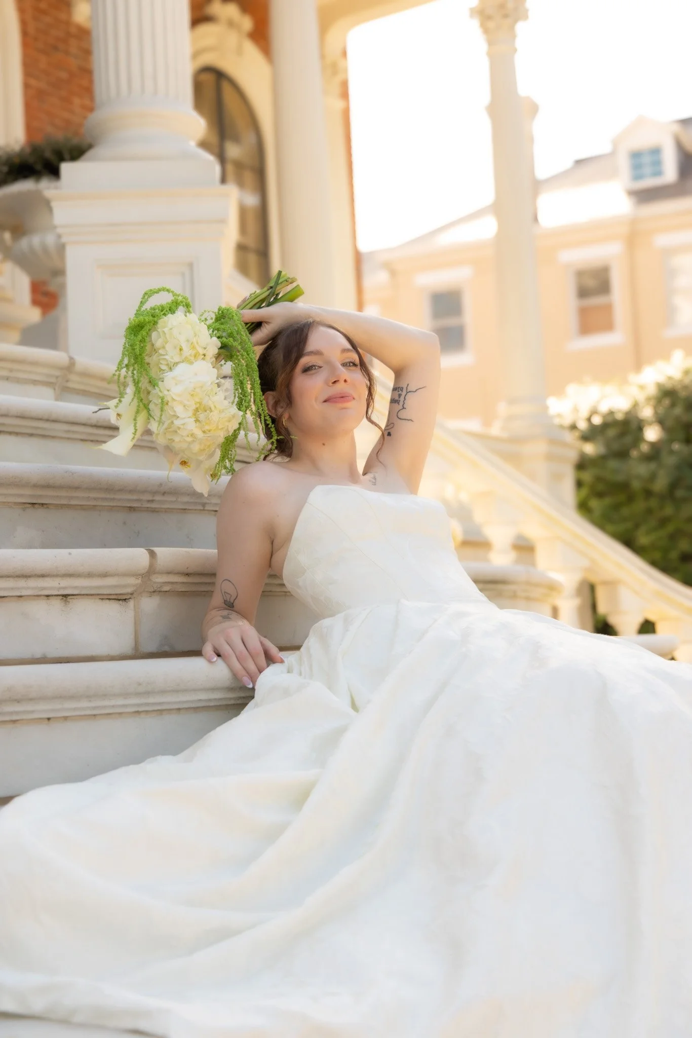 A woman in a wedding dress sitting on stairs outside a building, holding a bouquet of flowers, with her hand on her head and smiling.