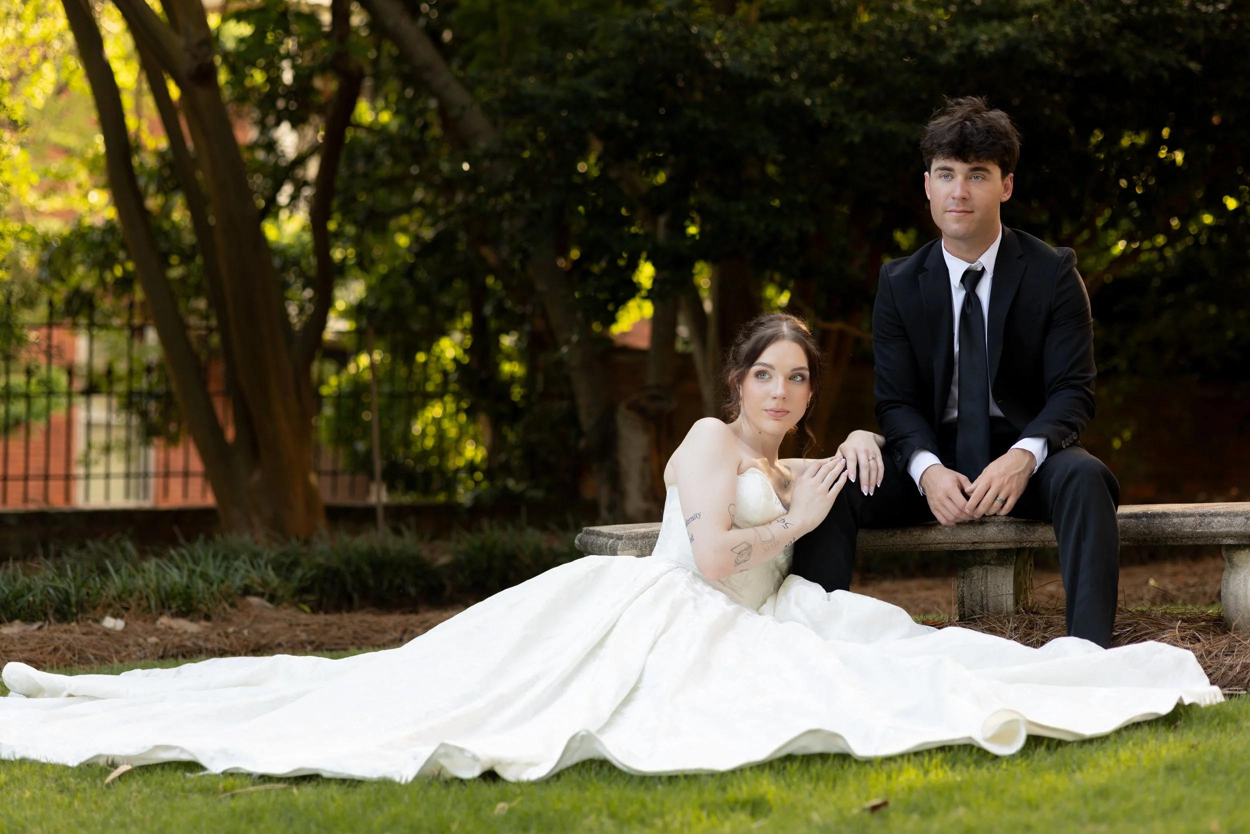 A bride sitting on a stone bench holding hands with a groom in a park. The bride is wearing a white wedding dress with tattoos on her arm, and the groom is dressed in a black suit with a white shirt and black tie. The background features trees and a 