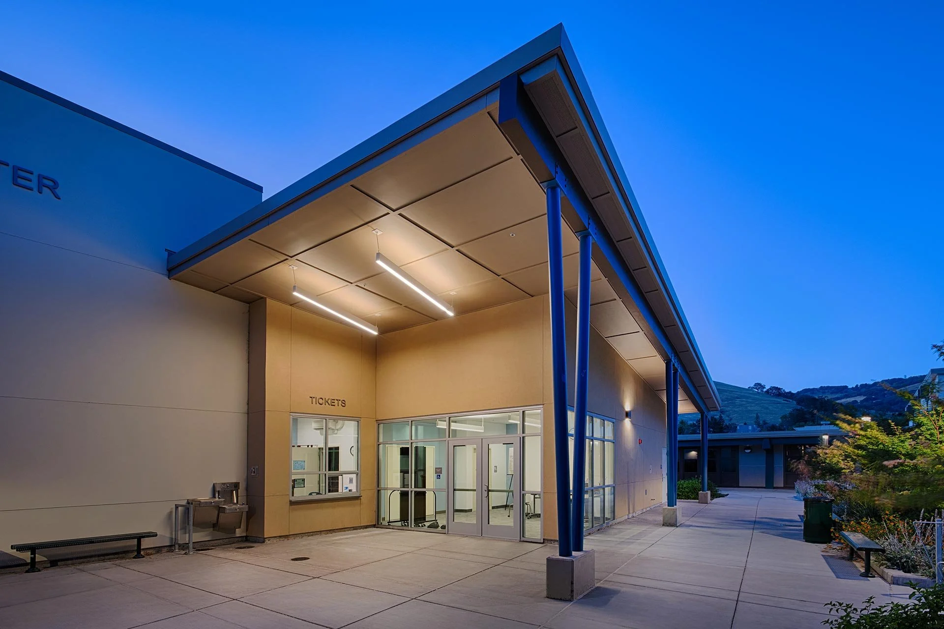 The Emily Gates Performing Arts Center entrance with ticket counter and glass doors, illuminated by overhead lights during dusk, with benches and greenery outside.