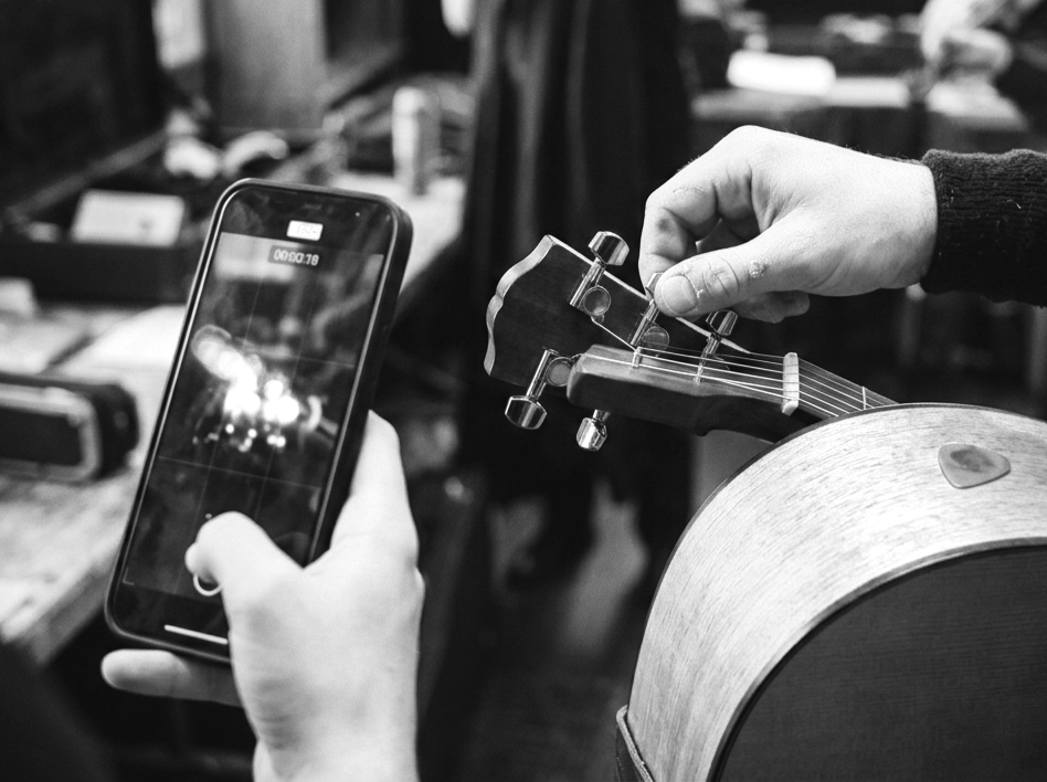 Person recording a video of a person tuning a guitar with a smartphone in a music workspace.